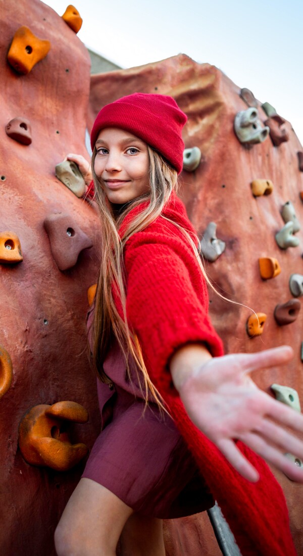 Jugendliche mit roter Mütze und Kleidung klettert an einer Kletterwand und streckt die Hand aus.
