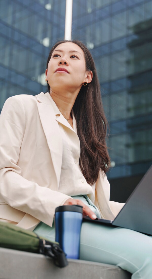 Eine junge Frau in Businesskleidung und Laptop auf dem Schoss vor einem großen Bürogebäude.