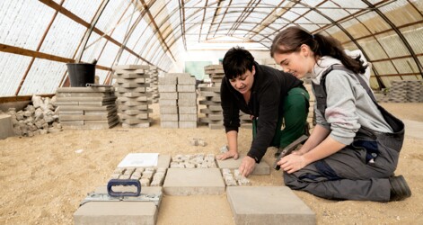 Zwei Frauen knien in einer Halle auf einem Sandboden. Die ältere Frau erklärt der jüngeren Frau, wie die Pflastersteine verlegt werden sollen. Im Hintergrund liegen Arbeitsmaterialien und Pflastersteine.
